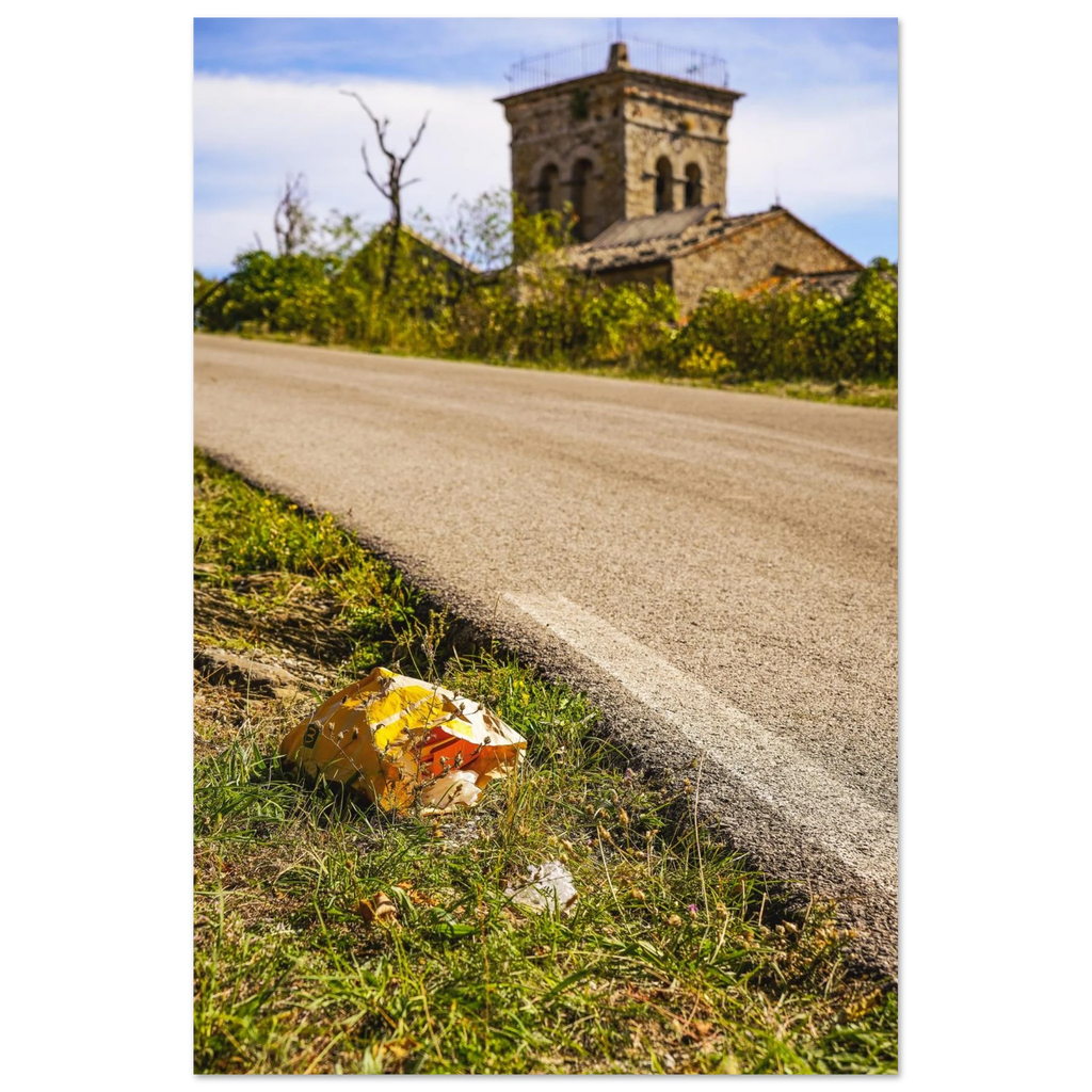 A discarded McDonalds bag sits in the grass beside a small road with a beautiful Italian church in the background.